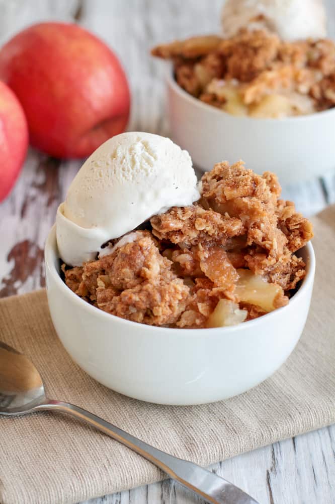 bowl of vegan apple crumble with ice cream on top of it with red apples in the background in a white bowl