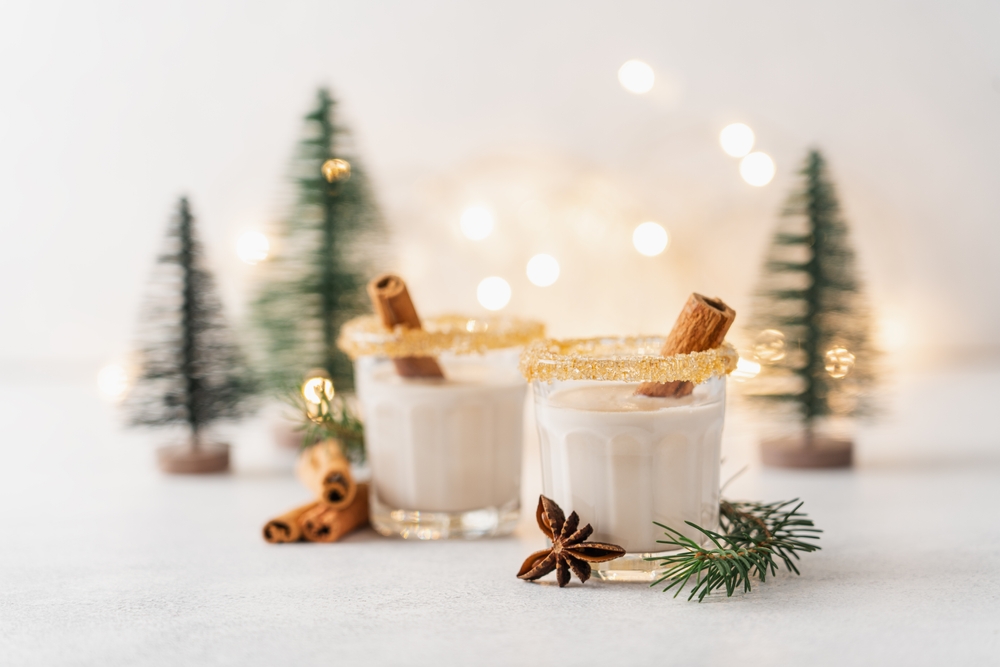 two gorgeous cups of vegan eggnog with sugar rims and with christmas trees in the background on a white surface