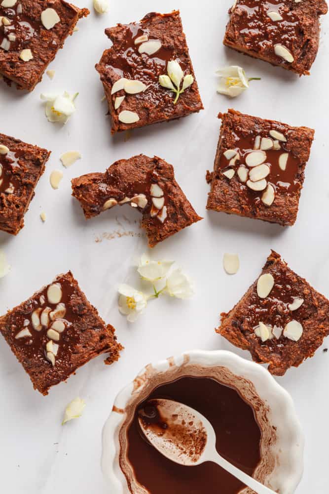 multiple vegan chocolate pumpkin brownies on the counter covered in toasted almonds and chocolate sauce with white flowers around 