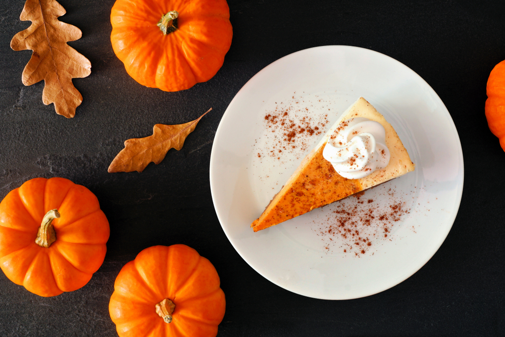 slice of orange non dairy pumpkin cheesecake with vegan whipped topping on a white plate with orange pumpkins and leaves around