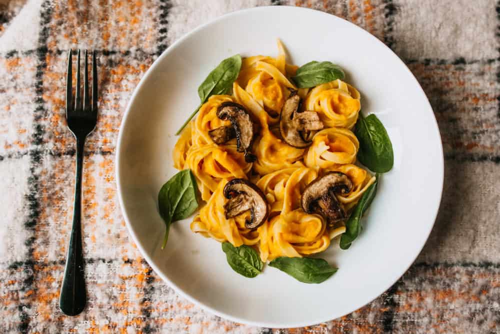 orange color yellow alfredo pasta with mushrooms and spinach on a plate with fork on checkered placemat