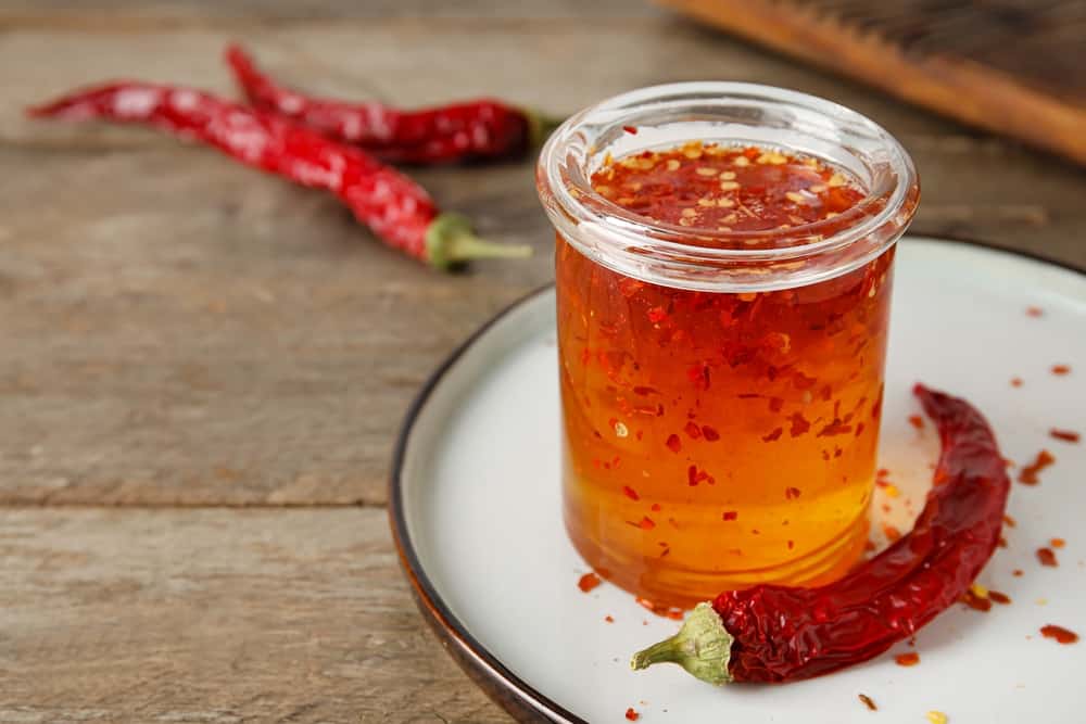 Jar of hot honey on a white plate with red dry chili peppers on a wooden table.