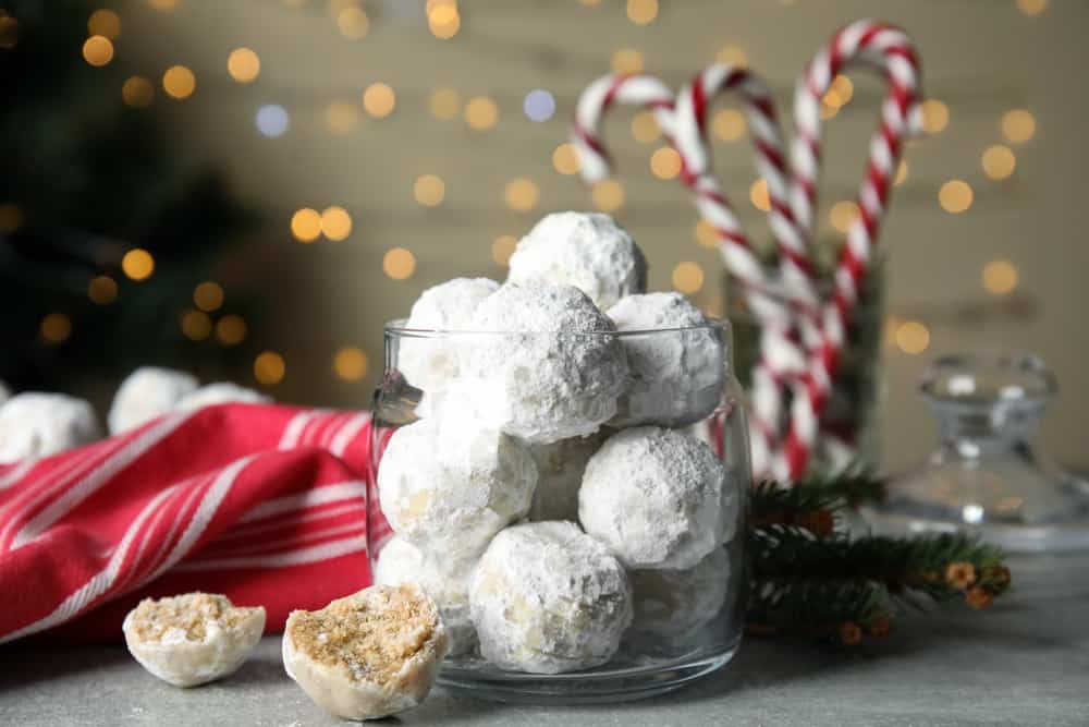 Glass jar with round butterball cookies covered in powdered sugar and one broken open on the counter. The background has strings of lights, a red cloth, and candy canes.