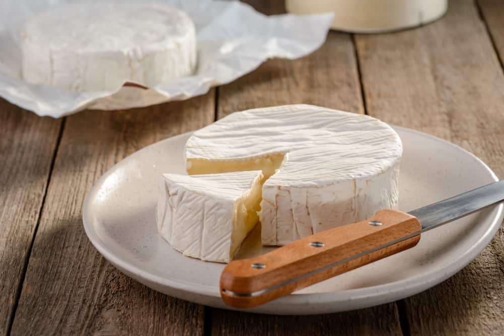 Wheel of brie cheese with a slice cut out on a white plate with a knife on a wooden table.