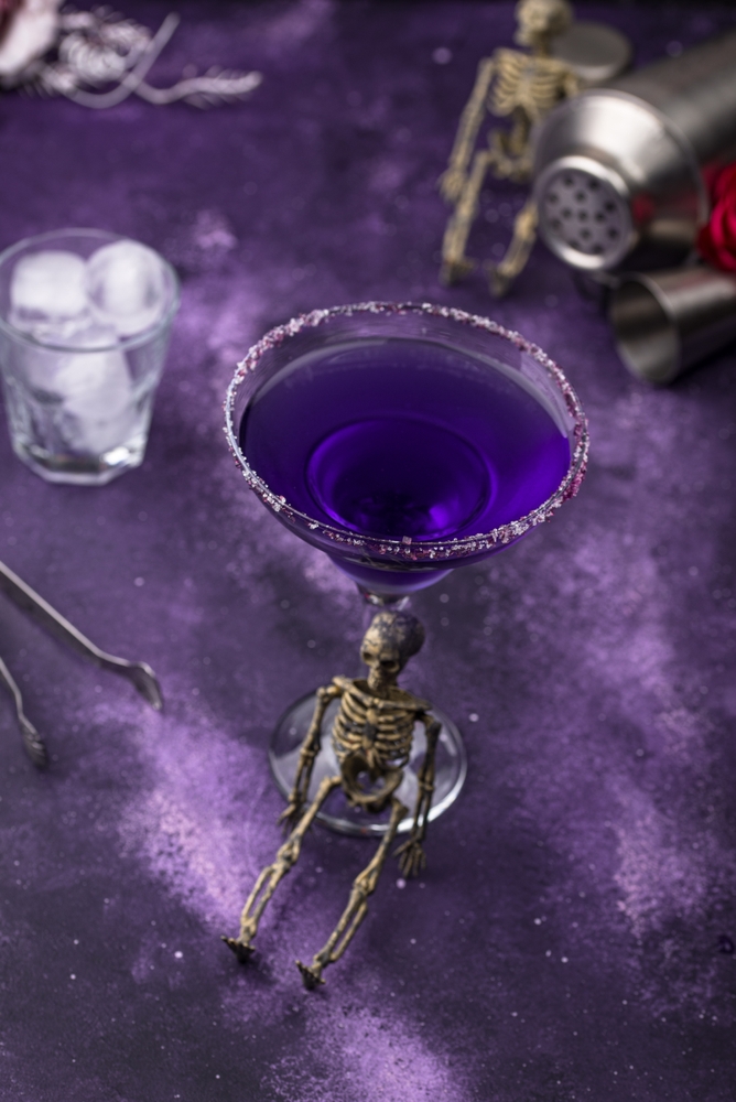 Looking down at a purple Halloween cocktail on a purple surface with small skeletons.