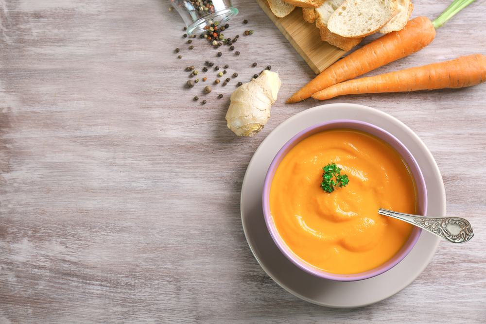 Flatlay photo of bowl of creamy carrot soup next to raw carrots, ginger, peppercorn, and bread on a gray wood table.