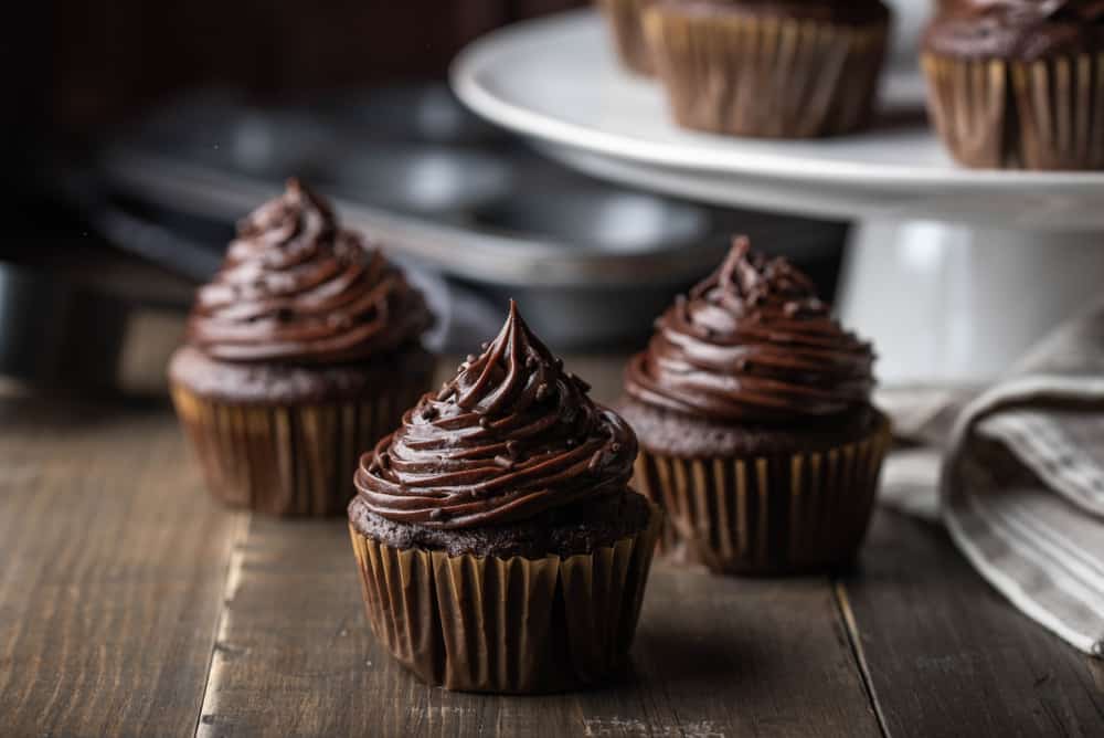 Three chocolate cupcakes on a dark wood table with more cupcakes on a stand in the back.