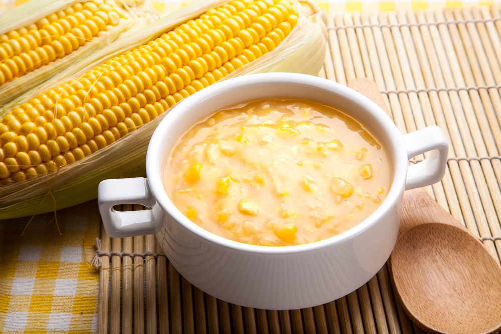 Creamy and thick corn soup in a white bowl next to two corn cobs.
