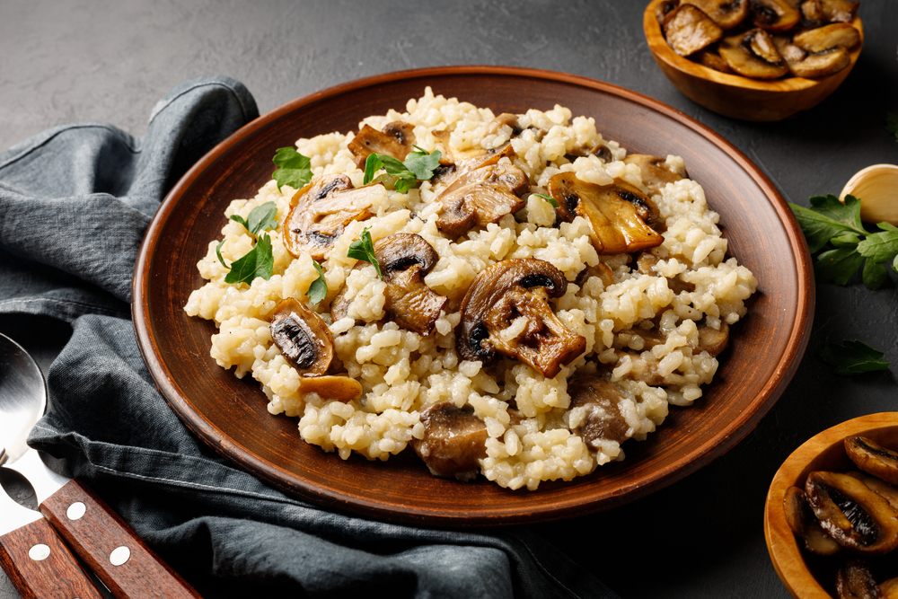 Vegan mushroom risotto in a wooden bowl next to a black napkin and bowls of sauteed mushrooms.