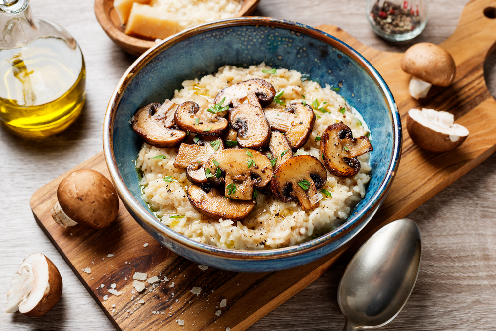Blue bowl of vegan risotto with sauteed mushrooms and seasonings on top on a cutting board surrounded by raw mushrooms a spoon and olive oil.