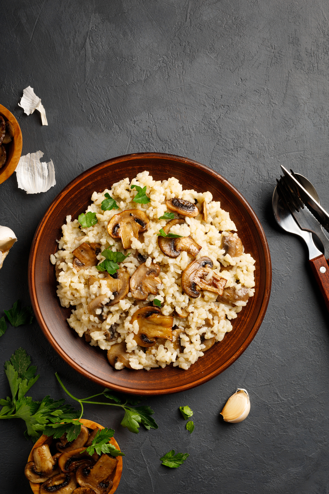 Brown bowl of vegan risotto topped with sauteed mushrooms and seasonings on a black background.