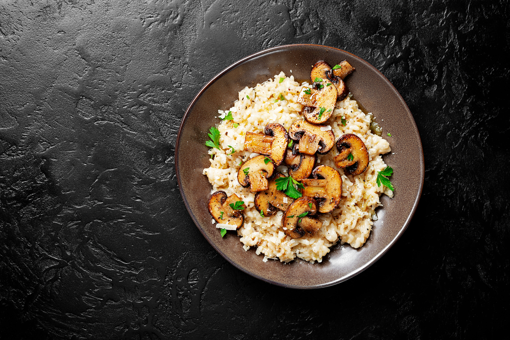 Flatlay of a brown plate of vegan risotto topped with sauteed mushrooms on a black background.