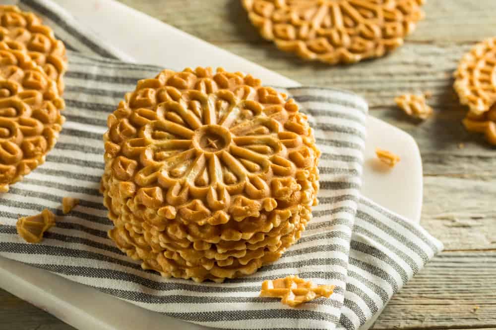 Stack of Vegan Pizzelles with intricate design on a grey and white striped towel on a wooden table.