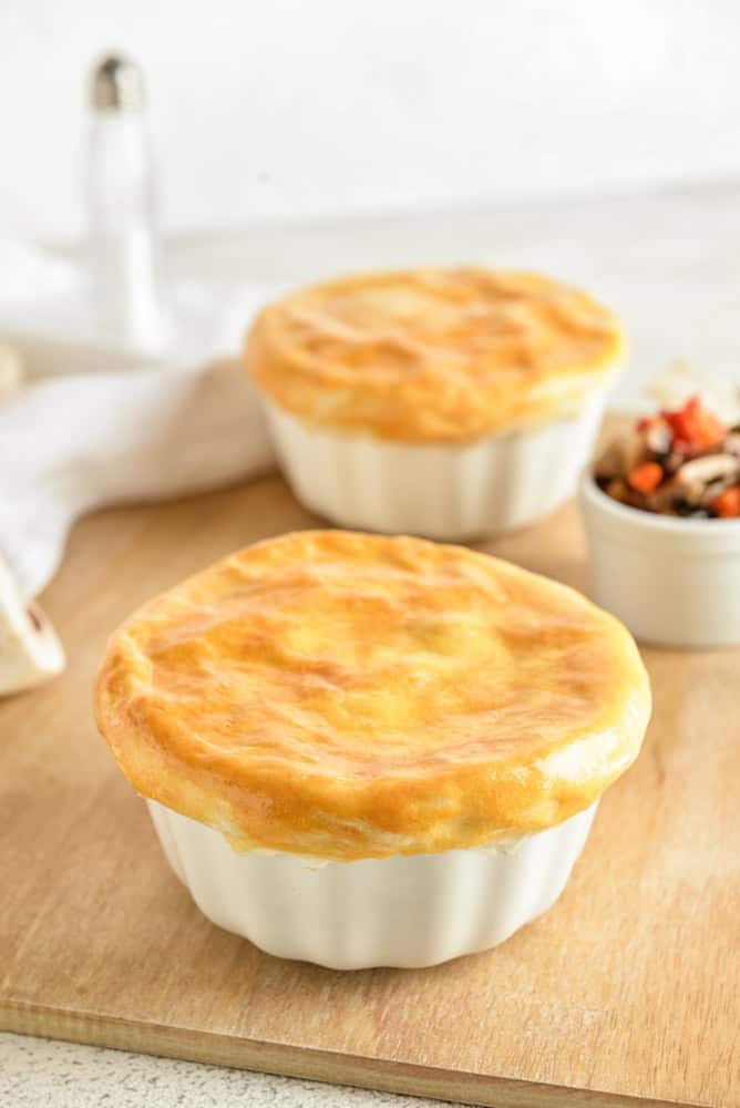 Two baked pot pies in white ramekins on a wooden cutting board.