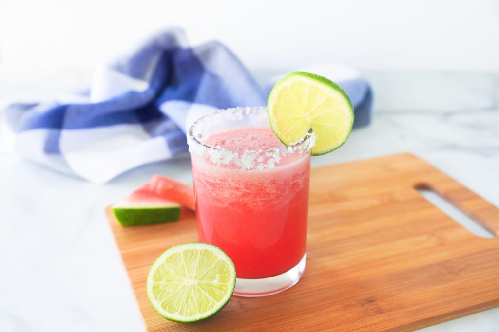 Glass of pink Frozen Watermelon Margarita with salted rim and lime garnish on a wooden cutting board next to a blue and white towel.