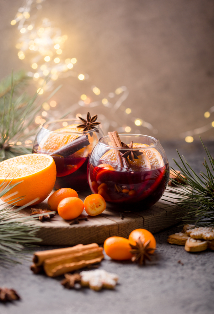 Two round glasses of mulled wine with fruit and cinnamon sticks on a serving tray with an orange. cranberries, and pine needles with Christmas lights in the background.