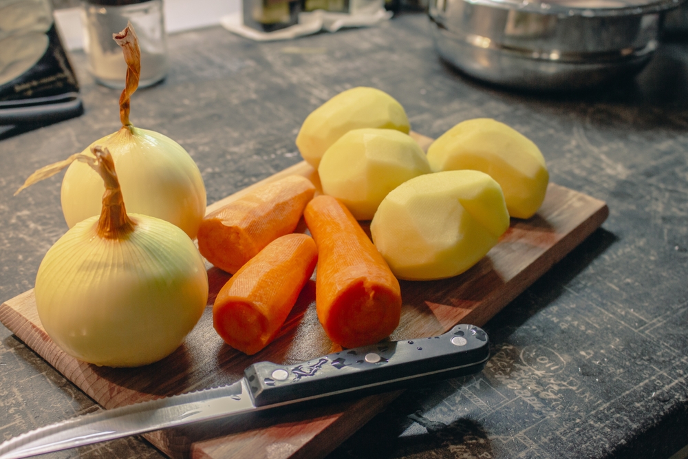 Peeled vegetables on wooden board with knife. Raw potatoes, onion and carrots on kitchen table. Preparing food concept. Vegetables on cutting board with kitchen knife. Soup ingredients. 