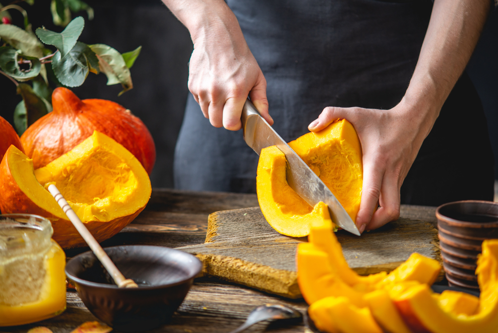 A woman cook cuts an orange pumpkin with a knife into slices for baking on a wooden table. Concept autumn food in a cozy dark kitchen with yellow flowers. Close up