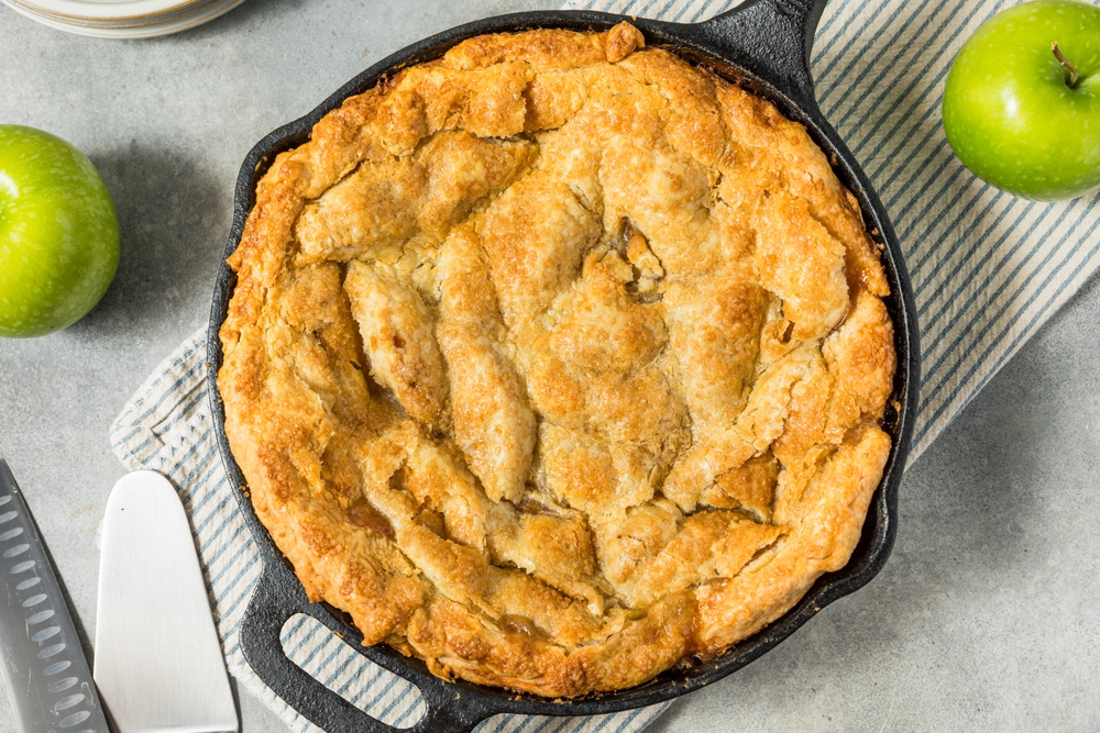 Pie cooked in a pan ona table with apples around it. 