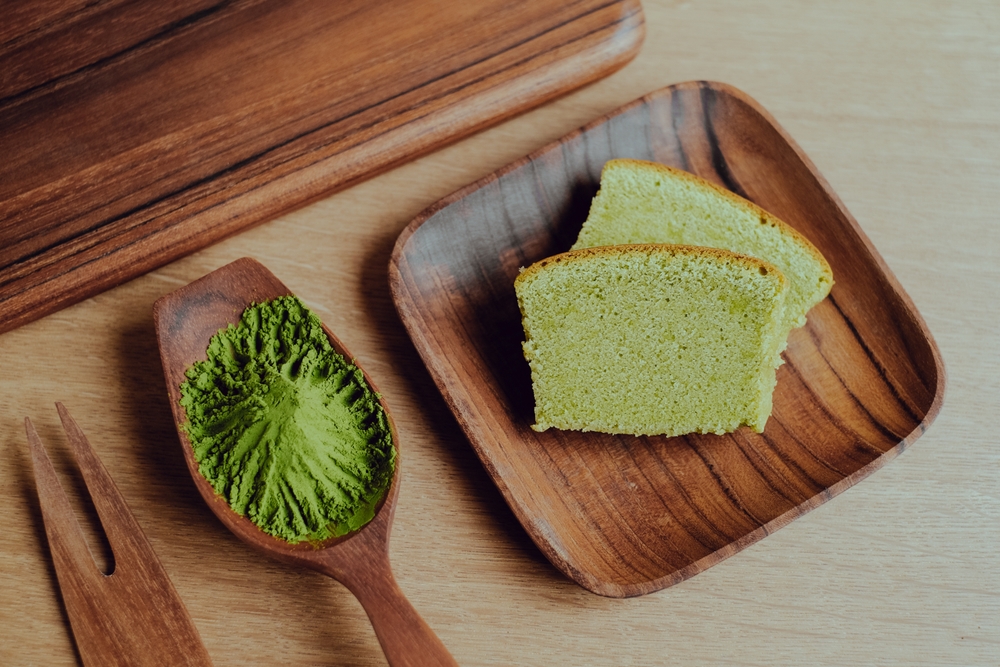 Two pieces of cake on a brown wood plate with a spoon full of matcha powder next to it. 