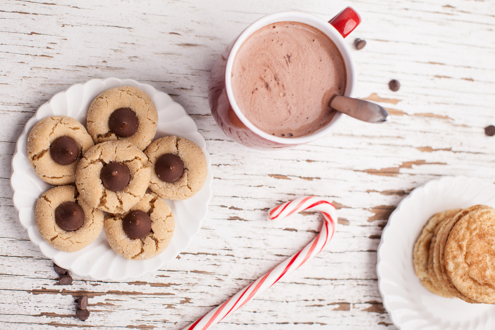Top horizontal view of plate of  cookies, hot chocolate, peppermint candy cane, and chocolate peanut blossom cookies on weathered white wood