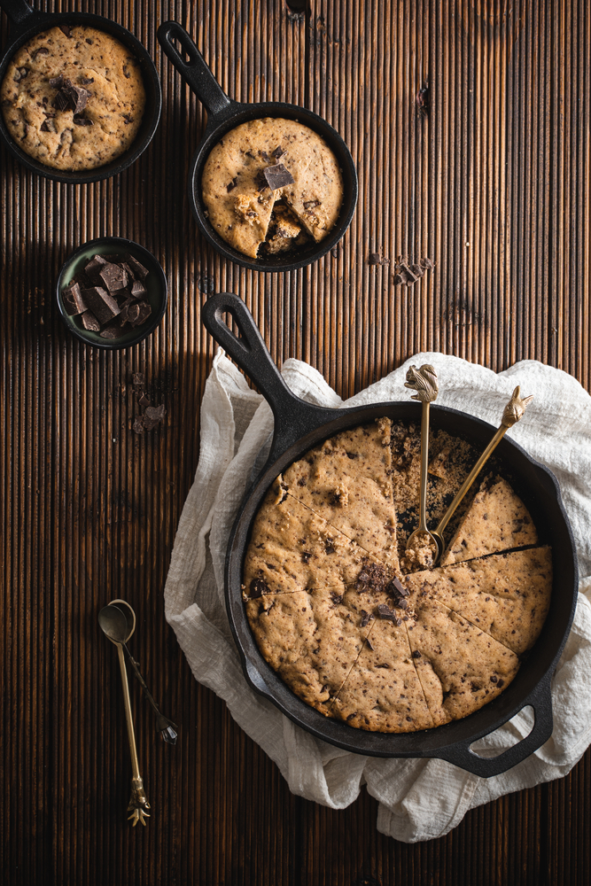 One chocolate chip cookie in a big black skillet on a kitchen towel and two small skillets with biscuits on a wooden table, a top view