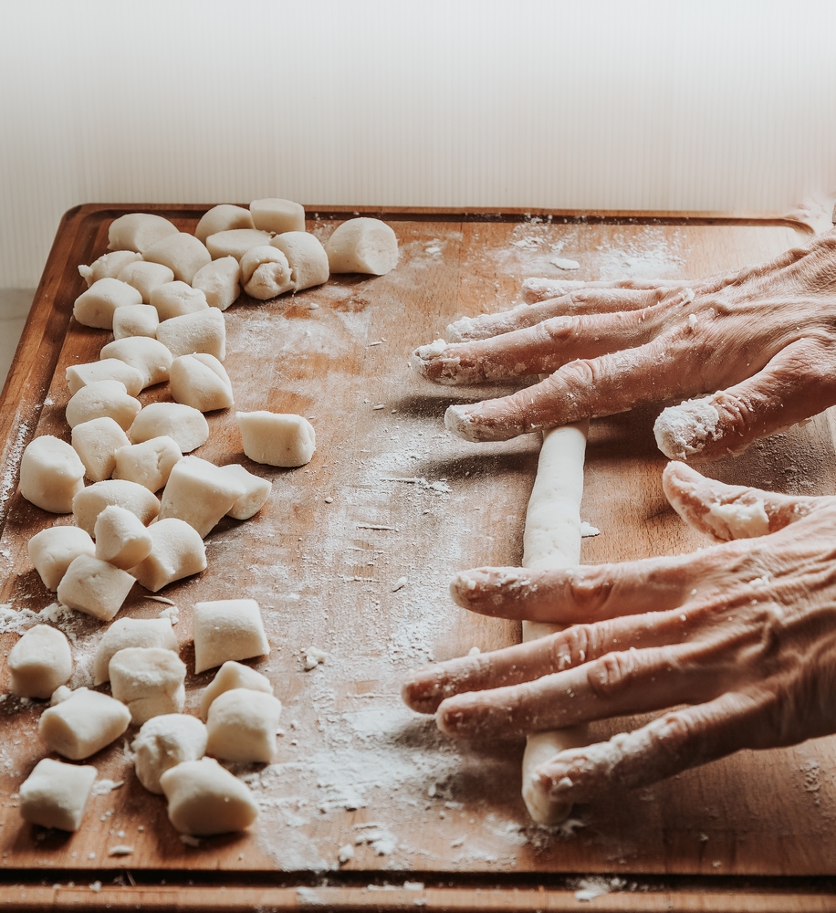 Image of the hands rolling out fresh dough for gnocchi pasta on a lightly floured wooden surface.