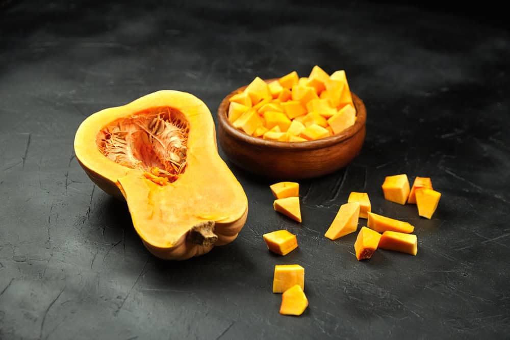 Chopped butternut squash, pumpkin half, fresh vegetable pieces in wooden bowl on black stone table, selective focus.