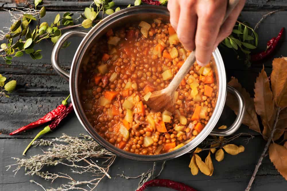high angle shot of a young caucasian man stirring lentils, with a wooden spoon, in a stainless steel cooking pan placed on a gray rustic wooden table
