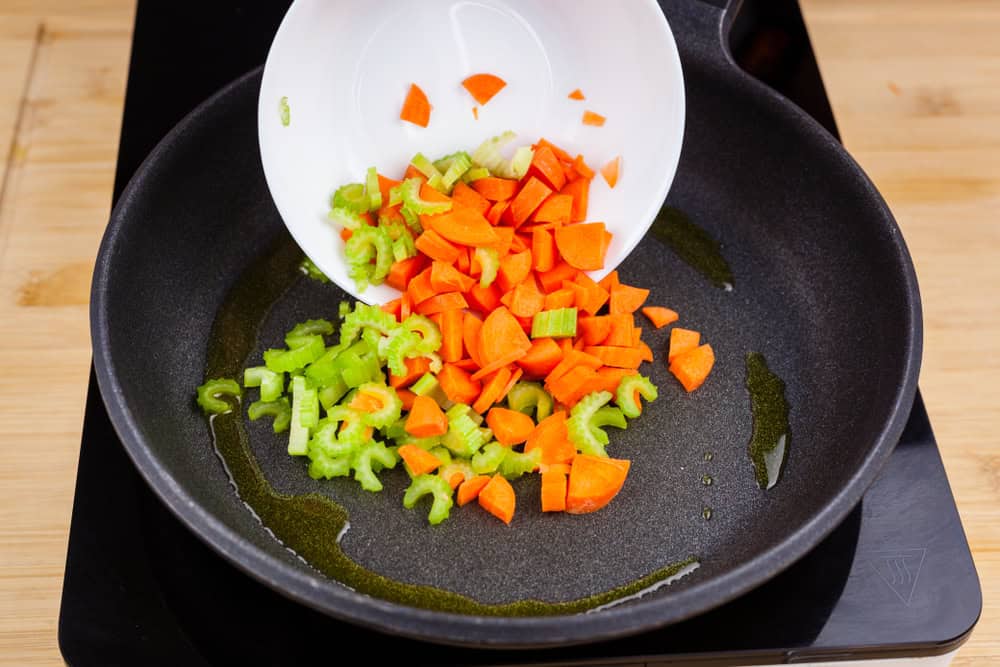 Hot frying pan with olive oil inside cooking while a bowl pours chopped celery and carrot dices on a ceramic hob with a wooden background.Vegan kitchen actions.