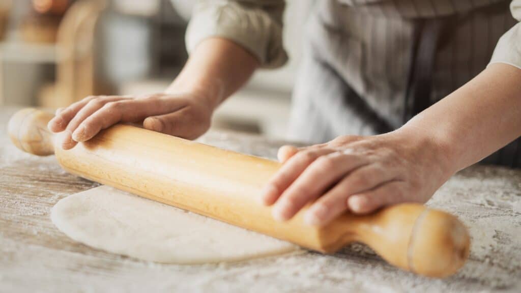 Kneading and rolling dough at the kitchen table. You can see the persons hands and the rolling pin. The article is a vegan ravioli recipe.